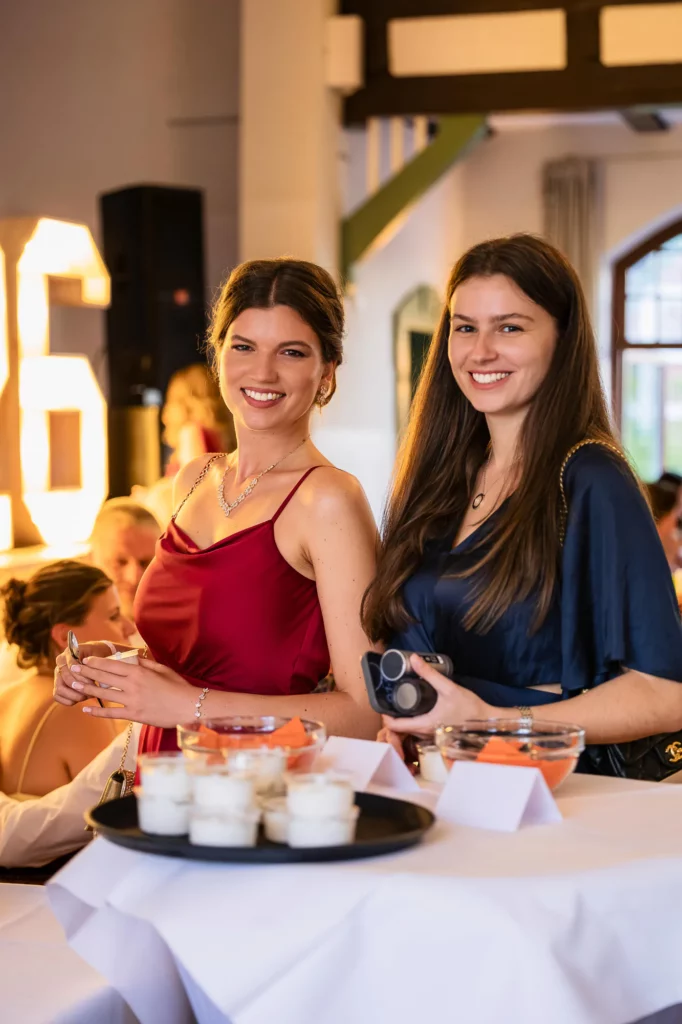 Fährhaus Kirschenland – Zwei junge Frauen in festlicher Abendgarderobe lächeln bei einem Abiball in die Kamera, im Hintergrund leuchtende Zahlen und gedeckte Stehtische. | Jannes Romund Fotografie
