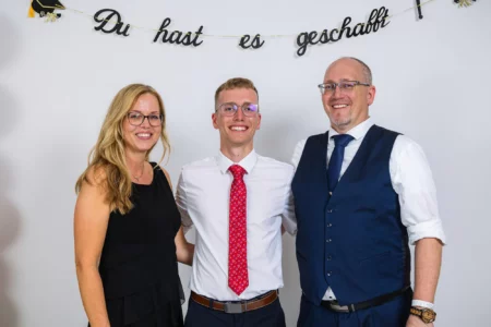 Fährhaus Kirschenland – Ein glücklicher Abiturient mit seinen Eltern vor einem weißen Hintergrund mit dem Schriftzug Du hast es geschafft bei einem Abiball in Stade. | Jannes Romund Fotografie
