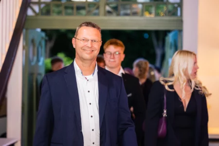 Fährhaus Kirschenland – Ein lächelnder Mann im dunkelblauen Anzug bei einem Abiball in Stade, im Hintergrund weitere Gäste in festlicher Abendgarderobe zu sehen. | Jannes Romund Fotografie