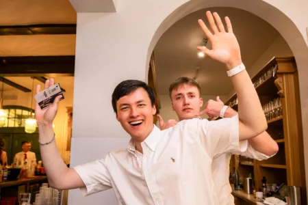 Fährhaus Kirschenland – Zwei junge Männer in weißen Hemden posieren fröhlich bei einem Abiball in Stade, einer hält eine Visitenkarte hoch, der andere winkt in die Kamera. | Jannes Romund Fotografie
