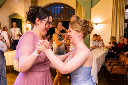 Fährhaus Kirschenland – Zwei lachende Abiturientinnen in festlichen Kleidern tanzen fröhlich miteinander auf einem Abiball in Stade bei einer Feier im Saal. | Jannes Romund Fotografie