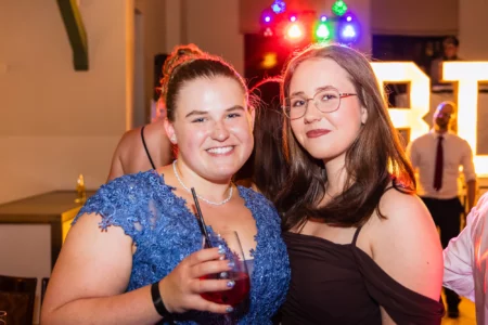 Fährhaus Kirschenland – Zwei junge Frauen in festlicher Abendgarderobe lächeln bei einem Abiball in Stade in die Kamera, im Hintergrund leuchtende Partybeleuchtung. | Jannes Romund Fotografie