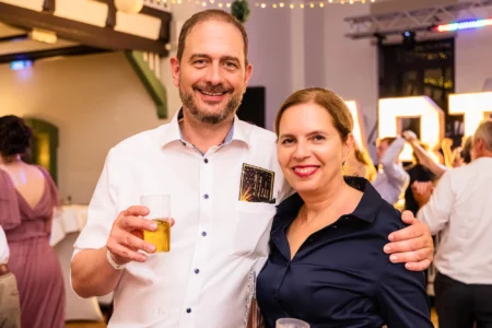 Fährhaus Kirschenland – Ein lächelndes Paar bei einem Abiball in Stade, der Mann hält ein Glas, im Hintergrund sind feiernde Gäste und leuchtende Buchstaben zu sehen. | Jannes Romund Fotografie