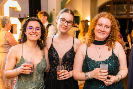 Fährhaus Kirschenland – Drei junge Frauen in eleganten Abendkleidern lächeln bei einem Abiball in Stade in die Kamera und halten Getränke in der Hand. | Jannes Romund Fotografie