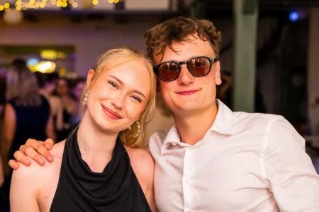 Fährhaus Kirschenland – Junges Paar lächelt bei einem Abiball in Stade in die Kamera, sie trägt ein schwarzes Kleid, er ein weißes Hemd und eine Sonnenbrille. | Jannes Romund Fotografie
