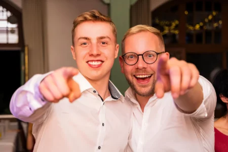 Fährhaus Kirschenland – Zwei fröhliche junge Männer in weißen Hemden zeigen bei einer Abiball-Feier in Stade direkt in die Kamera, ein Porträt voller Lebensfreude. | Jannes Romund Fotografie