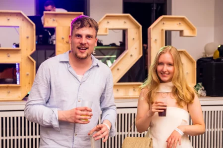 Fährhaus Kirschenland – Ein junges Paar hält Getränke vor großen, beleuchteten Holzbuchstaben bei einer Abiball-Feier in Stade für ein Porträtfoto in die Kamera. | Jannes Romund Fotografie