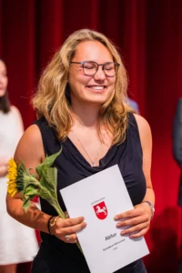 Abiballfotograf Stade – Eine glückliche Abiturientin hält ihr Abiturzeugnis und eine Sonnenblume in der Hand, während sie lächelt. | Jannes Romund Fotografie