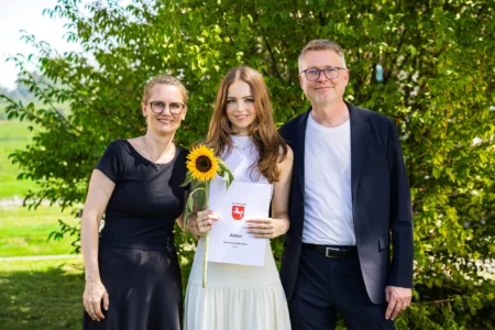 Abiballfotograf Stade – Eine junge Frau hält ihr Abiturzeugnis und eine Sonnenblume, umgeben von ihren Eltern in einer grünen Umgebung. | Jannes Romund Fotografie