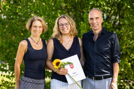 Abiballfotograf Stade – Eine junge Frau hält ihr Abiturzeugnis und eine Sonnenblume, umgeben von zwei Erwachsenen im Freien. | Jannes Romund Fotografie