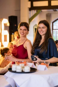 Fährhaus Kirschenland – Zwei junge Frauen in festlicher Abendgarderobe lächeln bei einem Abiball in die Kamera, im Hintergrund leuchtende Zahlen und gedeckte Stehtische. | Jannes Romund Fotografie