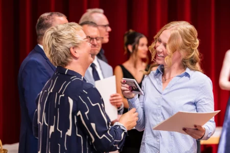 Abiballfotograf Stade – Zwei Frauen unterhalten sich fröhlich bei einer Veranstaltung, während andere Gäste im Hintergrund stehen. | Jannes Romund Fotografie