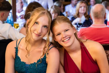 Fährhaus Kirschenland – Zwei junge Abiturientinnen lächeln in festlicher Abendgarderobe bei ihrem Abiball in die Kamera, im Hintergrund sind weitere Gäste zu sehen. | Jannes Romund Fotografie