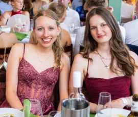 Fährhaus Kirschenland – Zwei junge Frauen in festlichen Abendkleidern lächeln bei einem Abiball in die Kamera, im Hintergrund weitere Gäste an festlich gedeckten Tischen. | Jannes Romund Fotografie