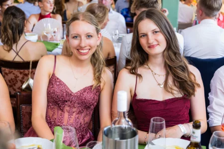 Fährhaus Kirschenland – Zwei junge Frauen in festlichen Abendkleidern lächeln bei einem Abiball in die Kamera, im Hintergrund weitere Gäste an festlich gedeckten Tischen. | Jannes Romund Fotografie