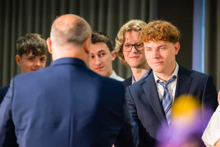 Abiballfotograf Stade – Junge Männer in Anzügen bei einer Zeremonie, einer schüttelt die Hand eines anderen, während die anderen zuschauen. | Jannes Romund Fotografie