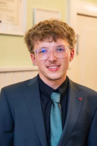 Fährhaus Kirschenland – Ein junger Abiturient mit Brille, schwarzem Hemd und grünem Schlips lächelt freundlich in die Kamera bei einem professionellen Fotoshooting. | Jannes Romund Fotografie