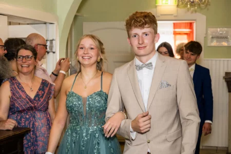 Fährhaus Kirschenland – Ein junges Paar in festlicher Abendgarderobe schreitet lächelnd bei einem Abiball durch einen Saal, begleitet von applaudierten Gästen im Hintergrund. | Jannes Romund Fotografie