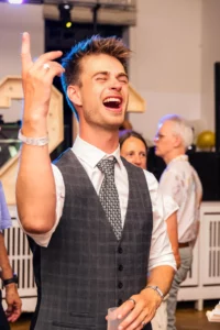 Fährhaus Kirschenland – Ein lachender Abiturient in festlicher Weste und Krawatte feiert ausgelassen auf einem Abiball in Stade mit erhobenem Arm und einem Glas. | Jannes Romund Fotografie