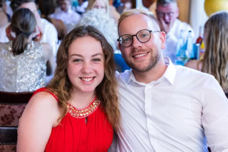 Fährhaus Kirschenland – Eine junge Frau im roten Abendkleid und ein Mann mit Brille lächeln gemeinsam bei einer festlichen Abiballfeier in die Kamera. | Jannes Romund Fotografie
