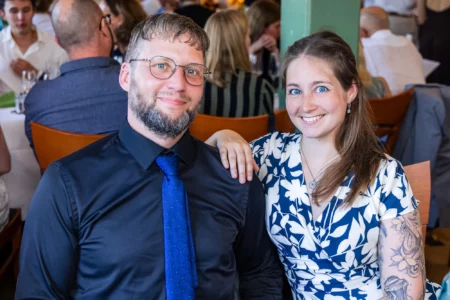 Fährhaus Kirschenland – Ein lächelndes Paar sitzt bei einem Abiball in Stade an einem Tisch; der Mann trägt Hemd und Krawatte, die Frau ein blau-weißes Sommerkleid. | Jannes Romund Fotografie