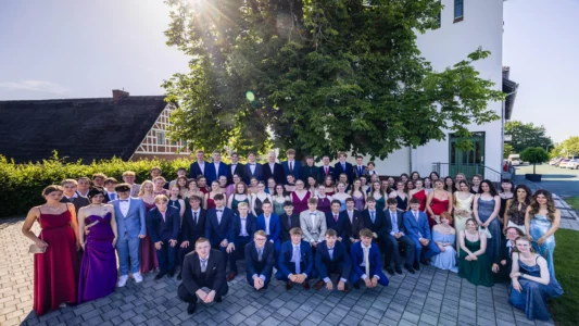Abiballfotograf Stade – Gruppenfoto von Schülern in eleganter Kleidung vor einem Gebäude, umgeben von Bäumen und sonnigem Wetter. | Jannes Romund Fotografie